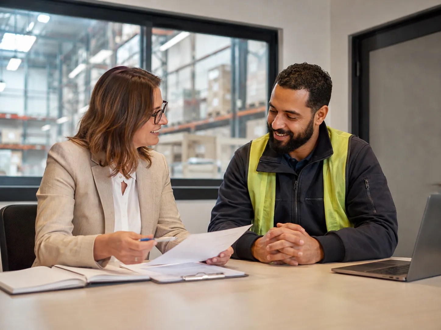 A workforce adviser reviews documents with a worker in a high-visibility vest at a desk overlooking a warehouse.